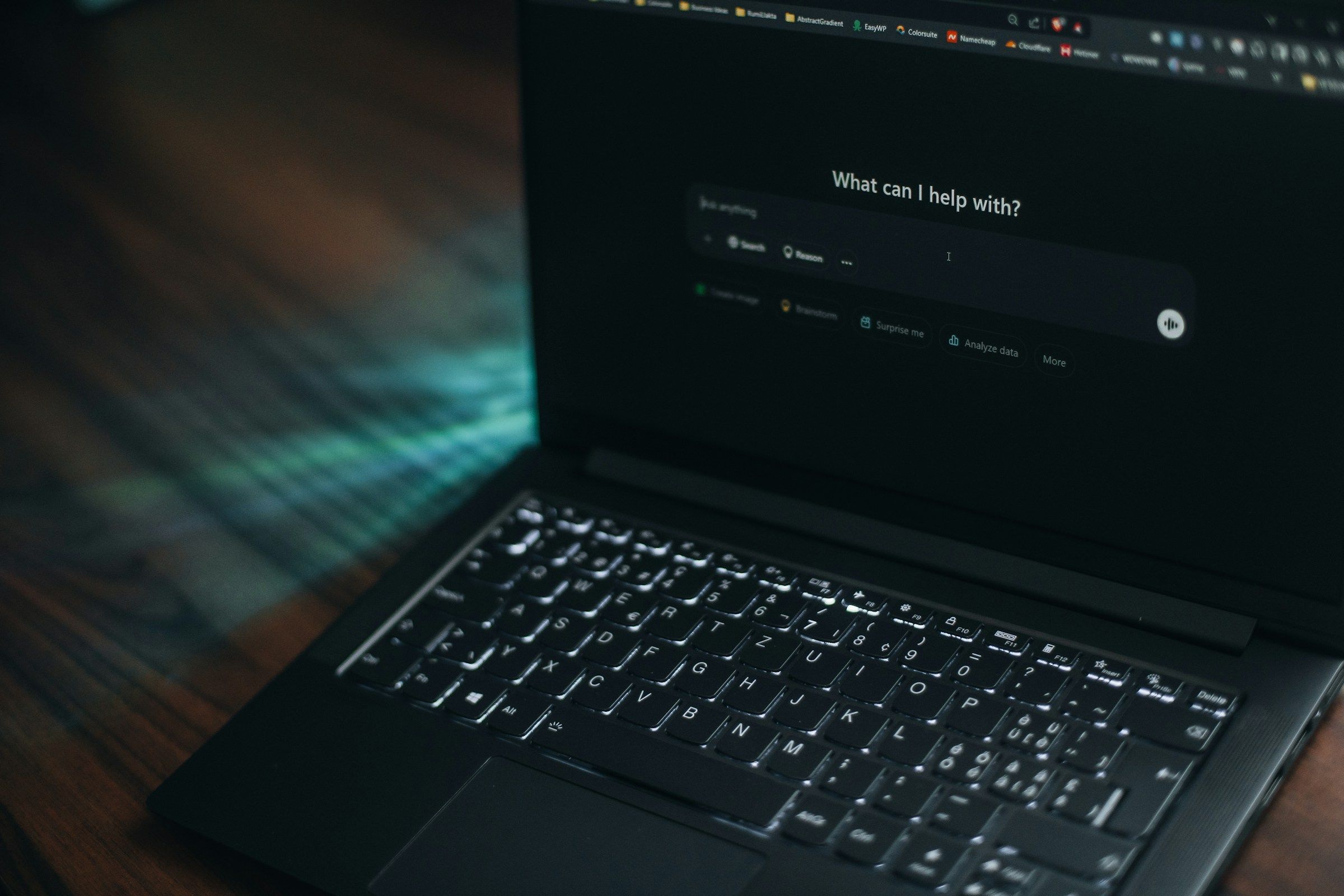 A laptop open on a wooden desk showing an AI assistant asking: What can I help with?